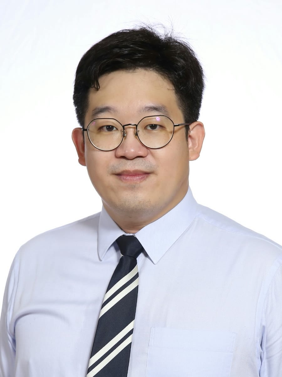 A man with round glasses, dark hair, wearing a light blue shirt and dark striped tie, against a white background.